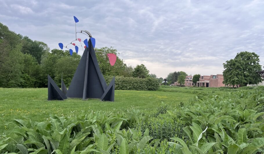 Sculpture moderne dans parc, ciel nuageux, bâtiments en arrière-plan.